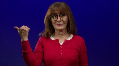 Kırmızı gömlekli bir kadın solu işaret ederek mavi arka planda neşeli hareketler sergiliyor..