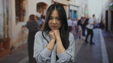 Genç bir kadın, hareketli bir sokakta, parlak bir günde çizgili gömleğiyle dalgın dalgın duruyor..