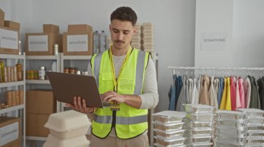 Yansıtıcı yelek giyen genç bir adam, kapalı bir odada dizüstü bilgisayar kullanırken kutularla ve kıyafetlerle çevrili bir hayır kurumunda bağış organize ediyor..