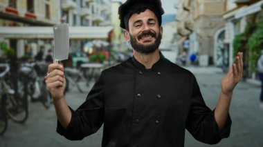 Sakallı, siyah üniformalı İspanyol aşçı balta tutuyor açık havada, hareketli bir sokakta gülümsüyor..