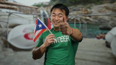 Deniz kenarında Puerto Riko bayrağı tutan genç bir adam baş parmağını kaldırmış, gönüllü gömleği giymiş, açık gökyüzü altında neşeli bir ifadeyle plaj manzarasının tadını çıkarıyor..