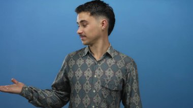 Genç İspanyol bir adam mavi arka plan duvarına karşı duruyor. Açık bir el hareketi ve desenli bir gömlek giyiyor..