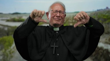 Açık hava parkı ortamında iki baş parmağıyla memnuniyetsizlik gösteren yaşlı rahip..