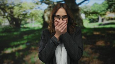 Parkta elleri ve ağzı kaplayan bir kadın dışarıda dururken canlı ağaçlarla çevrili, gözlüklü ve çizgili ceketli bir kadın şaşkınlığını dile getiriyor..