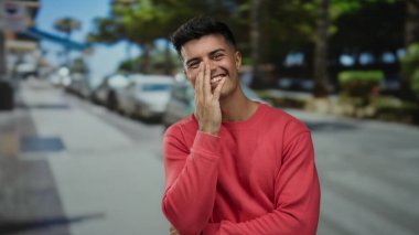 Kırmızı kazaklı genç adam güneşli bir şehir caddesinde gülümsüyor, rahat ve rahat bir atmosfer sergiliyor..