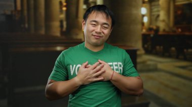 Gönüllü olarak etiketlenmiş yeşil gömlekli genç adam kilisede mutlu duruyor, ellerini göğsüne koyuyor, dini bir ortamda minnettarlık ve memnuniyeti ima ediyor..