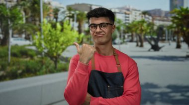 Kırmızı kazaklı ve gözlüklü genç bir adam şehir sokaklarında dikiliyor. Başparmağını kente odaklanmış ifadesiyle işaret ediyor..