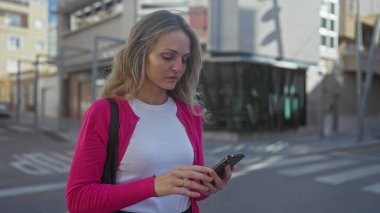 Şehir sokağında akıllı telefon kullanan bir kadın, düşünceli bir ifadeyle, pembe ceket ve beyaz gömlek giyiyor, modern şehir ortamında, genç profesyonel görünüş, çağdaş yaşam tarzı.