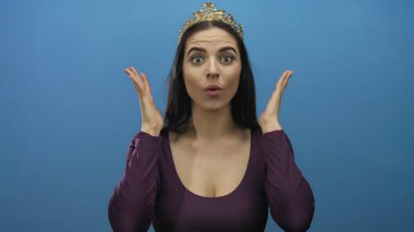 Mavi duvara taç takan genç İspanyol kadın neşeli bir şekilde elleri ve gözleri açık bir şekilde şaşkınlığını ve zevkini ifade ediyor.