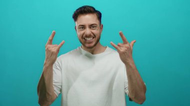 Sakallı genç adam kaya gibi el hareketleri yaparak canlı, mavi bir arka plana güvenerek gülümsüyor, enerjisini ve gençliğini gösteriyor..
