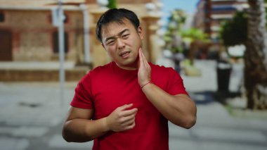 Kırmızı tişörtlü genç adam, şehir arka planında bulanık bir diş ağrısı çekiyor. İfadesinde acıdan rahatsız olduğunu ifade ediyor..