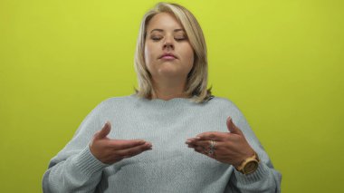 Sarışın ve gözleri kapalı bir kadın göğüs hizasında canlı yeşil stüdyo duvarına karşı avuç içi jest yapıyor; sakin denge farkındalığı.