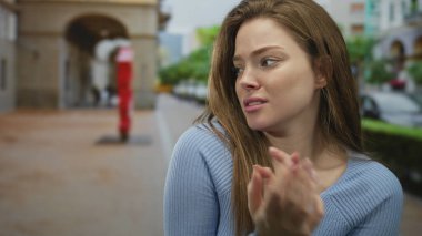 Bulanık arabaların ve sokak lambalarının arasında ellerini sıkan gergin ifadeye sahip genç beyaz kadın; endişe.