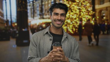 Gülümseyen sıradan adam, tatil ışıkları altında iki eliyle şehir caddesinde kahve fincanı tutuyor; memnuniyet.