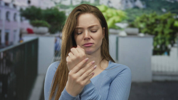 Женщина держит запястье одной рукой на размытом фасаде городской улицы при дневном свете; дискомфорт.