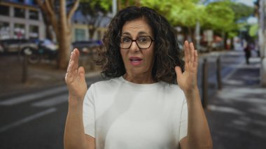 Orta yaşlı kıvırcık saçlı ve gözlüklü İspanyol kadın işlek cadde kavşağında ellerini ardına kadar açıyor..