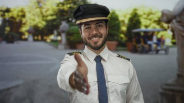 Sakallı ve yüzbaşı üniformalı genç adam gülümsüyor ve açık hava parkında arka planda bulanık ağaçlarla el ele tutuşuyor.