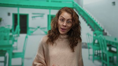 Restoran terasında, yeşil sandalyeli, şehir zemininde gözlüklü ve açık havada süveterli bir kadın şaşkınlığını dile getiriyor..