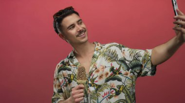 Çiçekli tişörtlü genç adam selfie çekiyor ve canlı kırmızı bir arka planda dondurma tutuyor neşeli tatil havası sergiliyor..