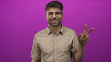 Adam mor stüdyoda iki eliyle barış işareti gösteriyor. Gözlük ve bej gömlek giyiyor. Zafer sevinci..