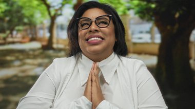 Parkta geniş bir şekilde gülümseyen bir kadın ağaçlarla çevrili, gözlük ve beyaz gömlek takan, mutluluk ve minnettarlık ifade eden, barışçıl neşeli atmosferi vurgulayan bir kadın..