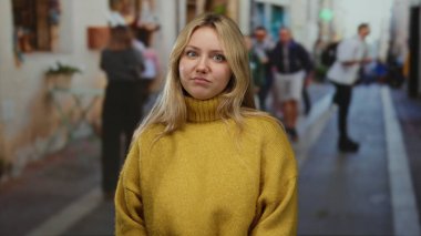 Yoğun şehir caddesinde dikilen sarı kazaklı bir kadın ve gündüz vakti arka planda yürüyen insanlar..