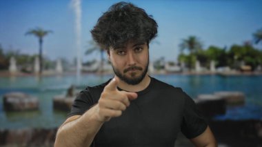 Parkta sakalları olan genç İspanyol bir adam parlak yeşilliğin arkasında düşünüyor ve işaret ediyor. Mavi gökyüzünün altındaki çeşme de tefekkür ve etkileşim öneriyor..