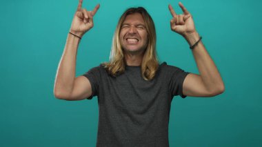 Sarışın, uzun saçlı, çıplak kollu sırıtan ve stüdyoda rock boynuzlarını gösteren bir adam..