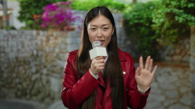 Kırmızı ceketli bir kadın mikrofon tutuyor şehir sokaklarında renkli çiçeklerle ve arka planda taş duvarlarla canlı şehir atmosferini yakalıyor..