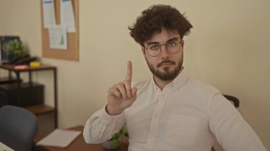 Ofis ortamında hayır işareti yapan, gözlüklü ve düğmeli tişörtlü İspanyol bir genç. Kapıda pano ve arka planda kağıtlarla oturuyor..