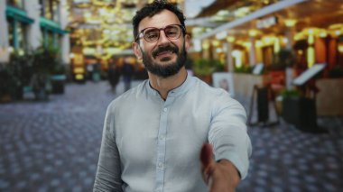 Gündüz vakti hareketli arka planı ve hareketli atmosferiyle canlı sokak sahnesinde elini uzatan sakallı İspanyol bir adam dostça etkileşim ve bağlantı sözü veriyor..