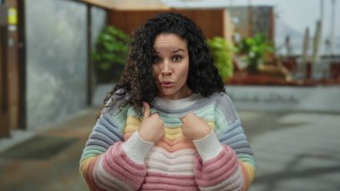 Renkli süveterli bir kadın dışarıda durup neşeli bir caddede kendini göstererek beklenmedik bir anı işaret ediyor..