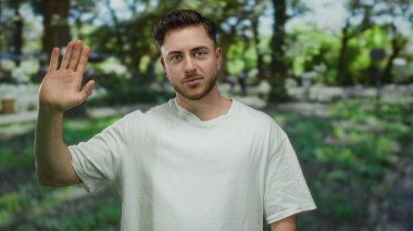 Beyaz tişörtlü genç adam güneşli bir günde yeşil ile çevrili bir parkta rahat bir ifadeyle kameraya bakıyor..