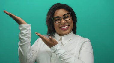 Gözlüklü ve beyaz gömlekli genç İspanyol kadın parlak bir şekilde gülümsüyor. Parlak yeşil bir arkaplan üzerinde ellerini uzatıyor. Olumlu ve coşkulu bir hava yayıyor..