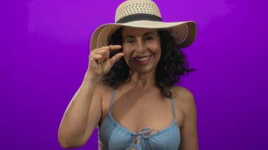 Mor arka planda şapkası olan, bikinili, orta yaşlı İspanyol bir kadın gülümsüyor..