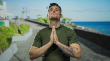 Zeytin rengi tişörtlü genç adam, deniz kenarında sakin sahiller ve okyanus arkaplanlarıyla meditasyon yapıyor, berrak gökyüzü altında huzurlu bir ifade ve sakinlik sergiliyor.
