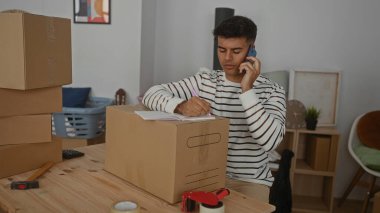 Çizgili süveter giyen genç adam oturma odasındaki karton kutuya yazarken telefonla konuşuyor. Etrafı mobilya ve malzemelerle çevrili..