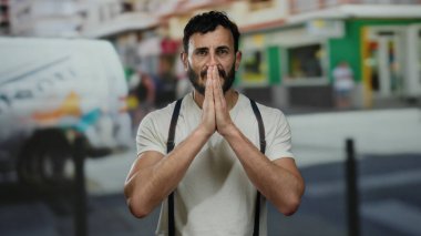 Sakallı İspanyol bir adam canlı bir sokakta namaz kılıyor. Pantolon askısı ve arka planda bulanık yayalar gibi beyaz bir gömlek giyiyor..