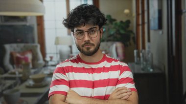 Çizgili gömlek giyen sakallı genç adam kollarını kavuşturup rahatlamış ama odaklanmış bir ruh hali yaratan iyi ışıklandırılmış bir otel salonunda güvenle duruyor..