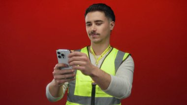 Yansıtıcı yelek giyen genç İspanyol adam, izole edilmiş kırmızı arka plana karşı telefon kullanıyor. Kasıtlı ifade ile şehir ortamında gönüllü çabayı temsil ediyor.