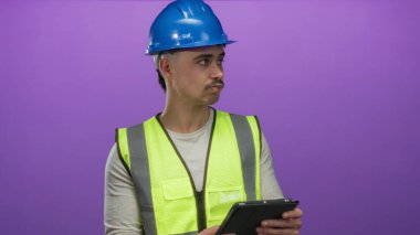 Yansıtıcı yelek ve şapka giyen genç adam, izole edilmiş mor arkaplan üzerinde bir tablet inceliyor, profesyonel odaklı teknoloji ve yapıyı sembolize ediyor..
