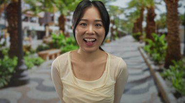 Canlı yeşil bir parkta palmiye ağaçlarıyla gülümseyen Çinli genç bir kadın, açık havada çekici ve neşeli bir ifade sergiliyor..