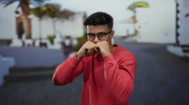 Kırmızı tişörtlü genç adam güneşli bir şehirde beyaz binalar ve palmiye ağaçlarıyla çevrili bir sokakta yumruklarıyla şakacı pozlar veriyor..
