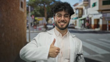 Sakallı, gözlüklü, beyaz önlüklü, güneşli bir şehir sokağında güven ve pozitiflik gösteren genç bir İspanyol..