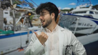 Sakallı, gözlüklü ve laboratuvar önlüklü genç adam liman zemininde gülümsüyor.