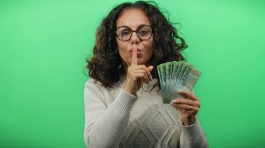 Elinde Rus rublesi taşıyan orta yaşlı bir kadın canlı yeşil bir arka plana karşı sessizlik işareti veriyor. Gizemli bir finans sahnesi yaratıyor..