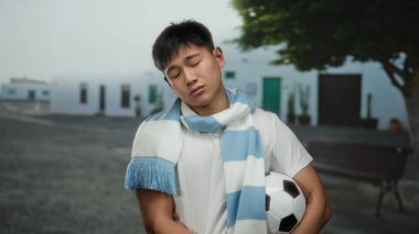 Beyaz gömlekli genç adam futbol topunu dışarıda tutarken aynı zamanda şehir caddesinde ağaca yaslanıyor ve gün boyunca sakin bir ifadeyle eşarp takıyor.