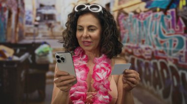 Bikini giyen ve Ley takan bir kadın akıllı telefon ve kredi kartını dışarıda duvar yazısının karşısında tutuyor..