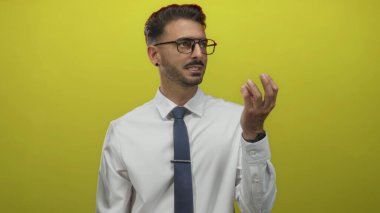 Gözlüklü, sakallı, beyaz gömlekli, mavi kravatlı, elini sarı stüdyoda kaldırmış bir adam..
