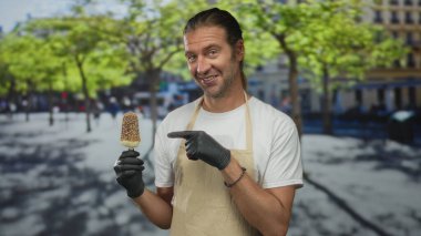 Sarı saçlı, önlüklü ve siyah eldivenli bir adam buzlu şeker tutuyor ve binayı işaret ediyor..